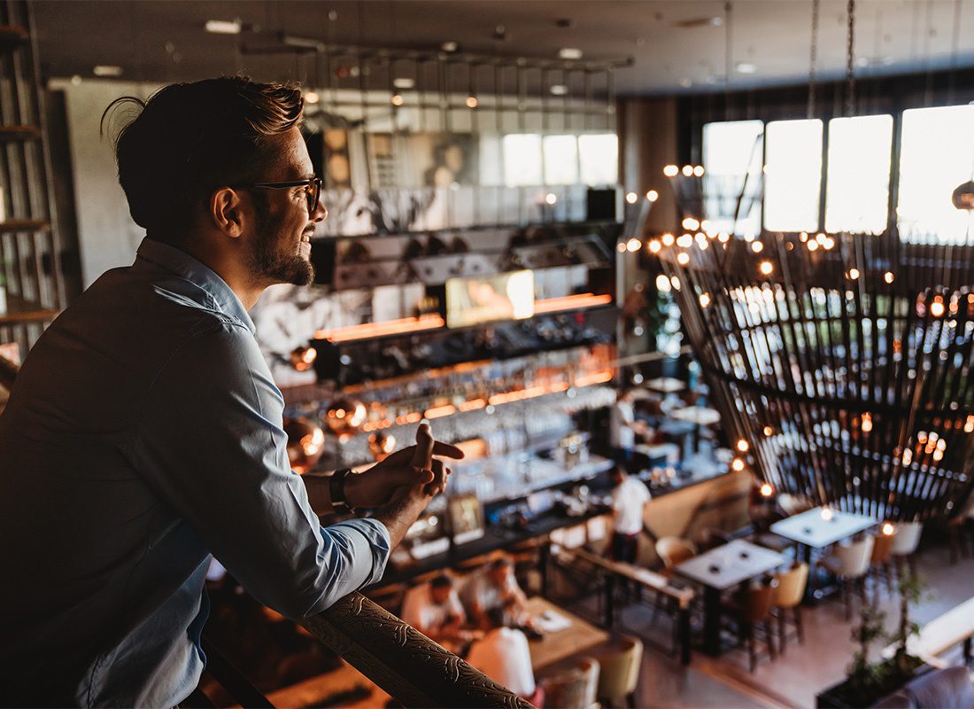 Business Insurance - Portrait of a Young Male Restaurant Owner Standing in the Loft Looking Down at the Busy Restaurant Area Below