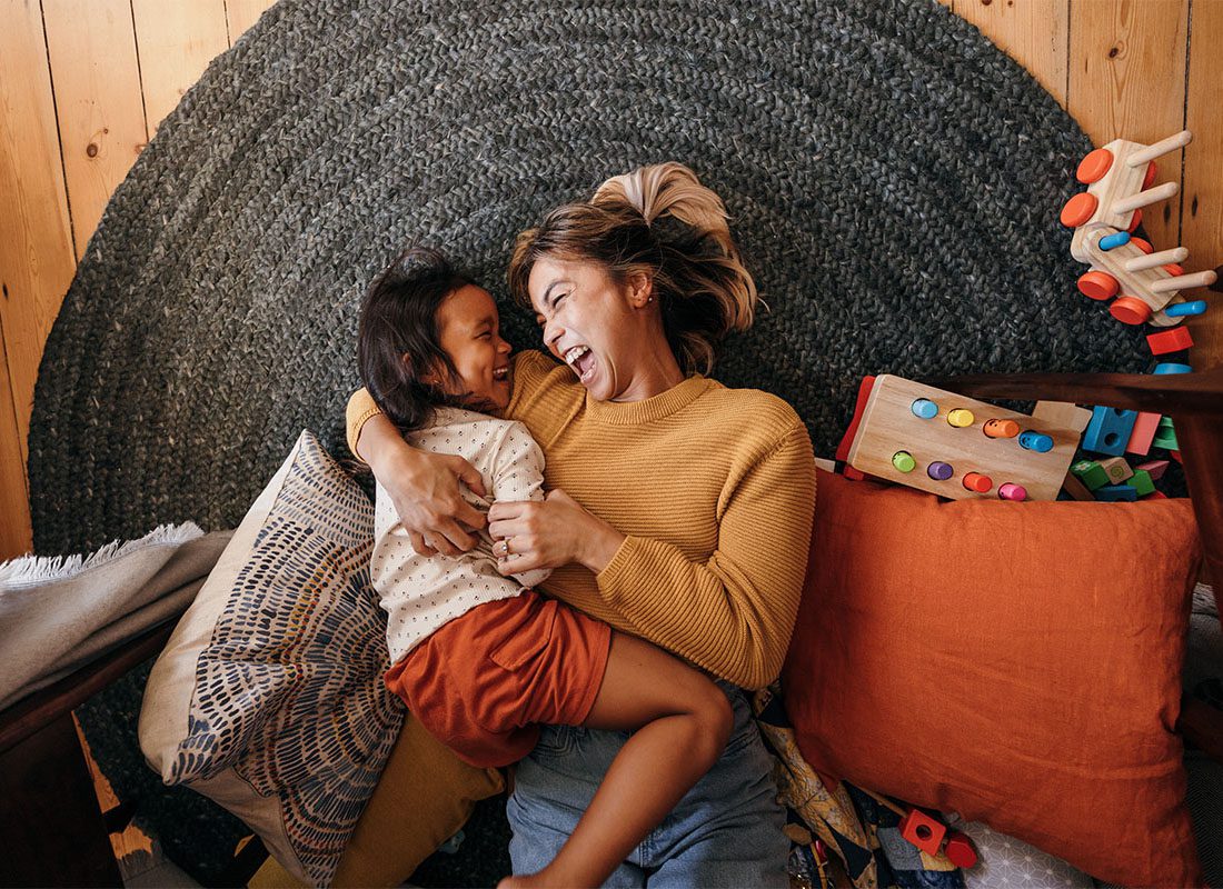 Personal Insurance - Portrait of an Excited Mother Having Fun Playing with her Young Daughter as They Both Lay on a Rug in the Living Room Surrounded by Pillows and Toys